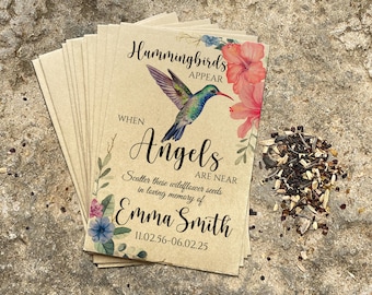May include: A stack of kraft paper seed packets with a hummingbird and floral design. Text reads "Hummingbirds Appear When Angels Are Near" and "Scatter these wildflower seeds in loving memory of Emma Smith". A pile of seeds is next to the packets.
