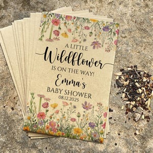 May include: A stack of kraft paper seed packets with a floral design and the text "A little Wildflower is on the way! Emma's Baby Shower 08.12.2025". A pile of seeds is next to the packets.