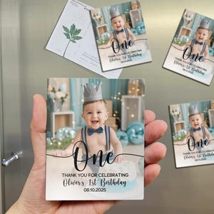 May include: Square magnets featuring a baby wearing a crown and bow tie, with the word "One" and the text "Thank You for Celebrating Oliver's 1st Birthday 08.10.2025". The magnets are held in a hand.