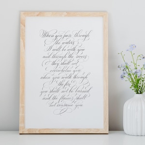 Può includere: Una stampa incorniciata con elegante calligrafia nera. Il testo recita: "Quando attraverserai le acque, io sarò con te..." La cornice è in legno chiaro, e un vaso bianco con fiori blu è a destra.