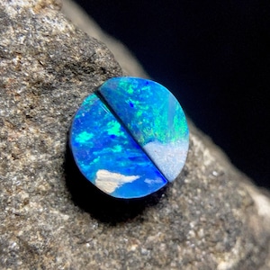 Puede incluir: Una piedra preciosa de ópalo pulida de forma circular con destellos vibrantes de color azul eléctrico y verde. La piedra está dividida por una fina línea diagonal oscura y muestra parches de material de matriz blanca en su superficie.