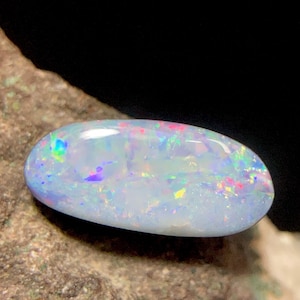 Puede incluir: Una piedra preciosa de ópalo en cabujón de forma ovalada con una base blanca translúcida y destellos vibrantes de color iridiscente, que incluyen reflejos de luz azul brillante, verde, amarillo y rojo en su superficie lisa, pulida y abovedada.