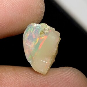 Könnte beinhalten: Ein roher Opal-Edelstein, der ein Farbenspiel mit roten, grünen und gelben Blitzen zeigt. Der Stein hat eine cremeweiße Grundfarbe und eine leicht unregelmäßige Form, gehalten zwischen Fingern vor dunklem Hintergrund.