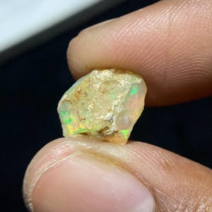 Pode incluir: Uma gema de opala bruta, não cortada, com uma cor de base amarelo creme. A opala exibe flashes de iridescência verde e amarela. A pedra é segurada entre os dedos, mostrando sua forma natural e bruta. Ideal para fazer joias ou colecionar.