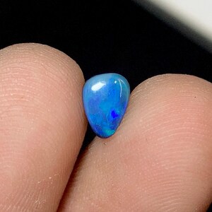 May include: A teardrop-shaped opal gemstone, displaying a vibrant blue color with internal flashes of green and darker blue. The polished surface reflects light, showcasing the stone's unique patterns and iridescence. The gemstone is held between two fingers.