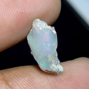 May include: A raw, uncut opal gemstone. The opal displays a milky white base color with flashes of iridescent green, red, and blue. The rough stone has a textured, natural appearance.