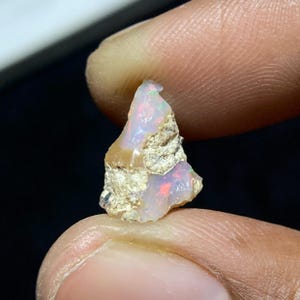 May include: A rough-cut opal gemstone with a mix of colors. The opal has a milky white base with flashes of red, green, and blue. The stone is held between two fingers, showcasing its natural, raw texture.
