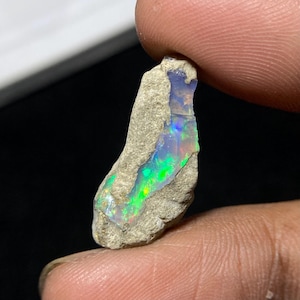 Op de afbeelding: Een ruwe, ongeslepen opaal edelsteen met levendige interne kleuren van groen, blauw en geel. De opaal is gezet in een lichtgrijze rotsmatrix, die zijn natuurlijke vorm laat zien. De edelsteen wordt tussen twee vingers gehouden.