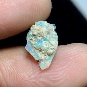 May include: A rough opal gemstone with a mix of colors, including blue, green, and red, is held between fingers. The opal has a textured, beige matrix and displays a play of light.