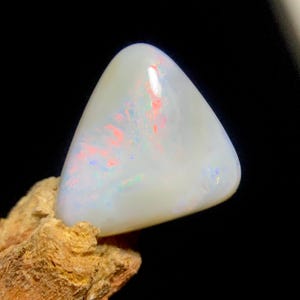 Op de afbeelding: Een driehoekige witte opaal edelsteen met een glad, gepolijst oppervlak met interne flitsen van rood, oranje en blauw licht. De edelsteen is geplaatst tegen een donkere achtergrond en rust op een gestructureerde, lichtbruine rotsachtige basis.