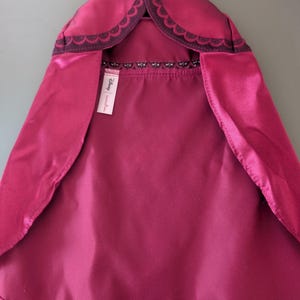 May include: A magenta cape with a scalloped dark purple trim around the hood. The cape has a satin-like finish and a tag with the Disney brand name.