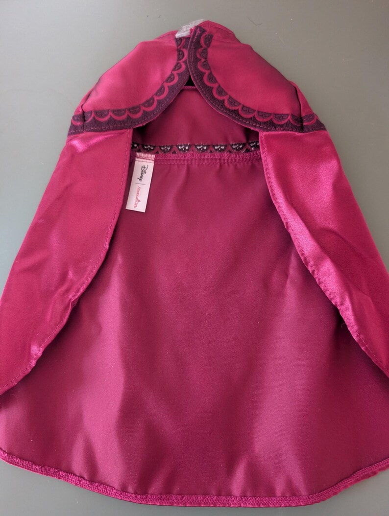 May include: A magenta cape with a scalloped dark purple trim around the hood. The cape has a satin-like finish and a tag with the Disney brand name.