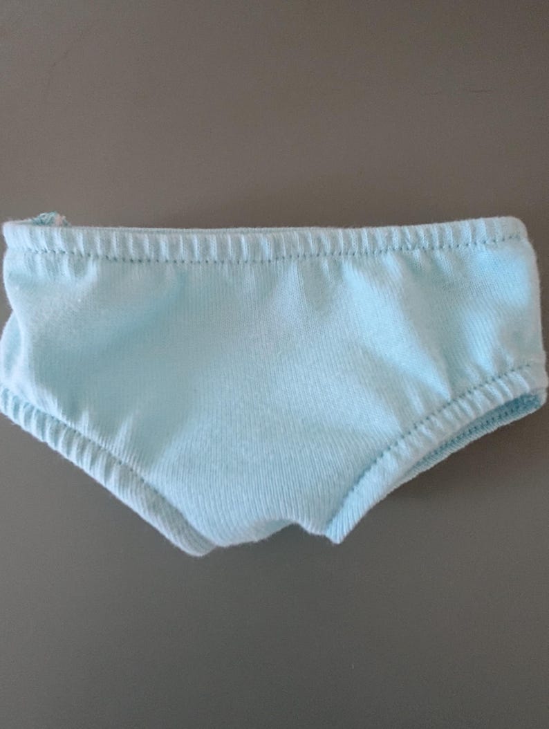 May include: Light blue underwear with elastic waistband and leg openings. The garment is made of a soft, light-colored fabric. The underwear is designed with a classic brief style.