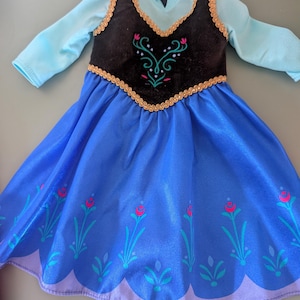 May include: A doll dress featuring a blue skirt with floral accents and a black bodice with gold trim. The dress has long sleeves and a high neckline. The bodice has an embroidered design with pink and green accents.