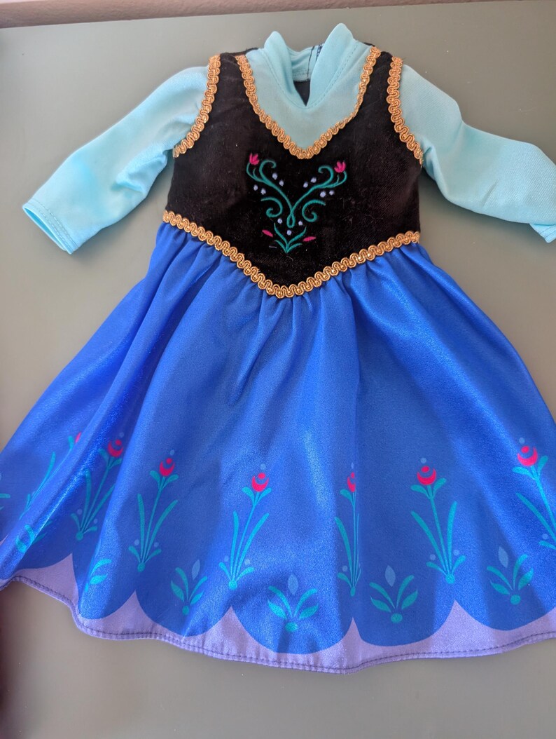 May include: A doll dress featuring a blue skirt with floral accents and a black bodice with gold trim. The dress has long sleeves and a high neckline. The bodice has an embroidered design with pink and green accents.