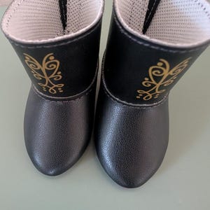 May include: A pair of black doll boots with gold decorative accents. The boots have a textured exterior and a white interior lining. The design includes a gold floral pattern on the side of each boot.