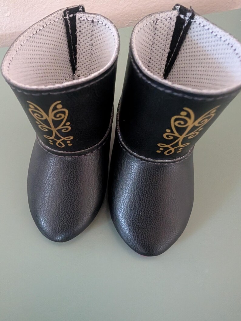 May include: A pair of black doll boots with gold decorative accents. The boots have a textured exterior and a white interior lining. The design includes a gold floral pattern on the side of each boot.