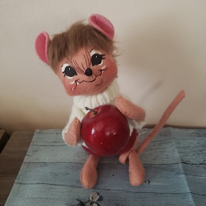 May include: A brown and pink felt mouse doll wearing a white sweater and holding a red apple.