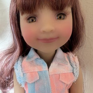 May include: A doll with brown eyes and long eyelashes, wearing a pink and blue tie-dye vest. The doll has long, auburn hair and rosy cheeks. The vest has two pockets and a red tag with white text.