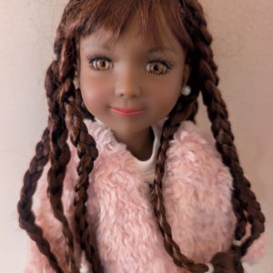 May include: A doll with brown skin, brown eyes, and long, braided brown hair. The doll is wearing a pink fuzzy jacket over a white top. The doll has a slight smile and is wearing pearl earrings. The doll is a child's toy.