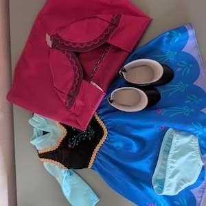 May include: A doll outfit featuring a blue dress with a black bodice, a pink cape, black shoes, and light blue underwear. The dress has gold trim and a floral design. The cape has a scalloped edge.