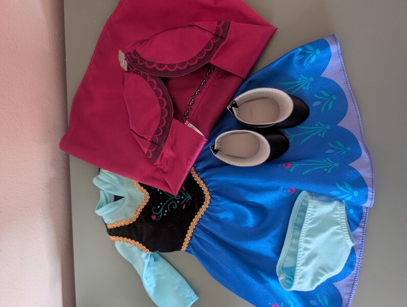 May include: A doll outfit featuring a blue dress with a black bodice, a pink cape, black shoes, and light blue underwear. The dress has gold trim and a floral design. The cape has a scalloped edge.