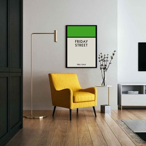 May include: A black framed print with a green rectangle at the top and the text "FRIDAY STREET" in black. The text "PRO.DAH" is in black at the bottom of the print. The print is hanging on a white wall above a yellow armchair.