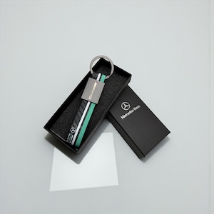 May include: A Mercedes-Benz AMG keyring with a black, white, and teal strap, presented in a black gift box. The keyring features a metal tag with "AMG" and a silver ring. The box is open, revealing the keyring against a dark interior.