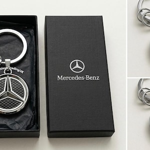 Puede incluir: Un llavero plateado de Mercedes-Benz con un emblema redondo y un llavero, presentado en una caja de regalo negra. La caja tiene el logotipo y el texto de Mercedes-Benz. Se muestran llaveros adicionales con superficies grabadas.