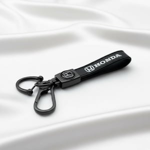 Op de afbeelding: Zwarte sleutelhanger met een Honda-logo en een metalen sluiting. De sleutelhanger heeft een zwarte band met het woord "HONDA" in witte letters. De sluiting is matzwart.