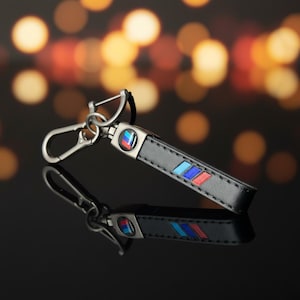 May include: Black leather keychain with a silver clasp and a colorful emblem. The keychain features a rectangular design with blue, red, and light blue stripes. The background is blurred with warm, out-of-focus lights.