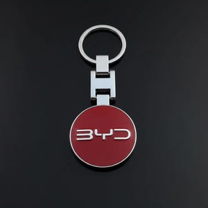 May include: A silver metal keychain with a circular red emblem. The emblem features the letters "BYD" in silver. The keychain has a ring for keys and a rectangular connector.