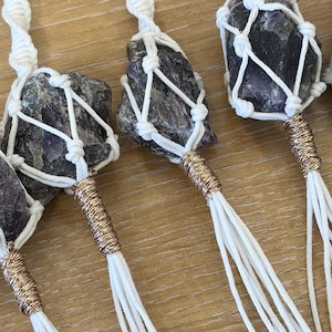 May include: Close-up of several amethyst crystal keychains. Each keychain features a raw, purple and gray amethyst stone encased in a white macrame net, with a gold-colored metal accent and white tassels.