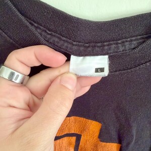 May include: A black t-shirt with a crew neckline and a white interior tag featuring the letter L in a black square. The fabric shows a faded texture, and a portion of an orange graphic design is visible at the bottom of the frame.