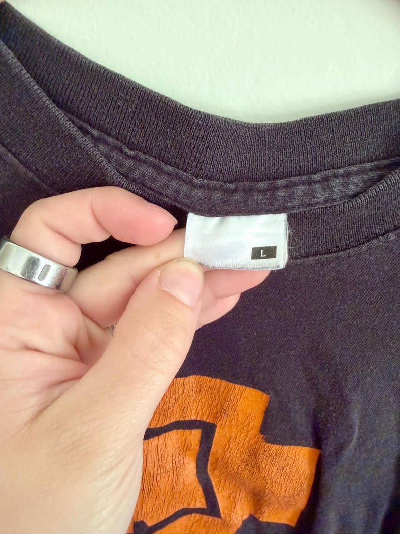 May include: A black t-shirt with a crew neckline and a white interior tag featuring the letter L in a black square. The fabric shows a faded texture, and a portion of an orange graphic design is visible at the bottom of the frame.