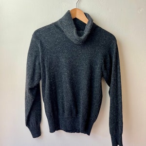 Könnte beinhalten: Ein dunkelgrauer Rollkragenpullover hängt an einem Holzbügel vor einer weißen Wand. Der Pullover hat lange Ärmel und einen gerippten Rollkragen. Der Stoff wirkt weich und leicht strukturiert.