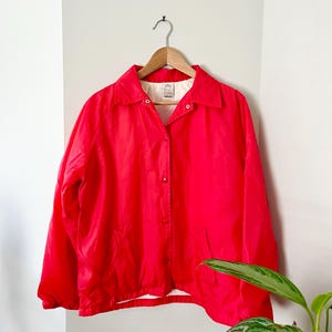 May include: A vibrant red jacket with a collared neckline and button-up front hangs on a wooden hanger. The jacket has long sleeves and two pockets. The jacket is made of a lightweight material.