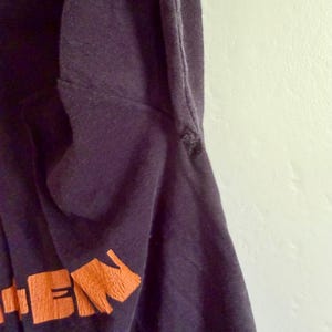 May include: A dark navy blue t-shirt featuring a portion of orange text printed on the lower section. The fabric shows a visible seam along the side and a small frayed area near the armhole, with a smooth texture throughout the garment.