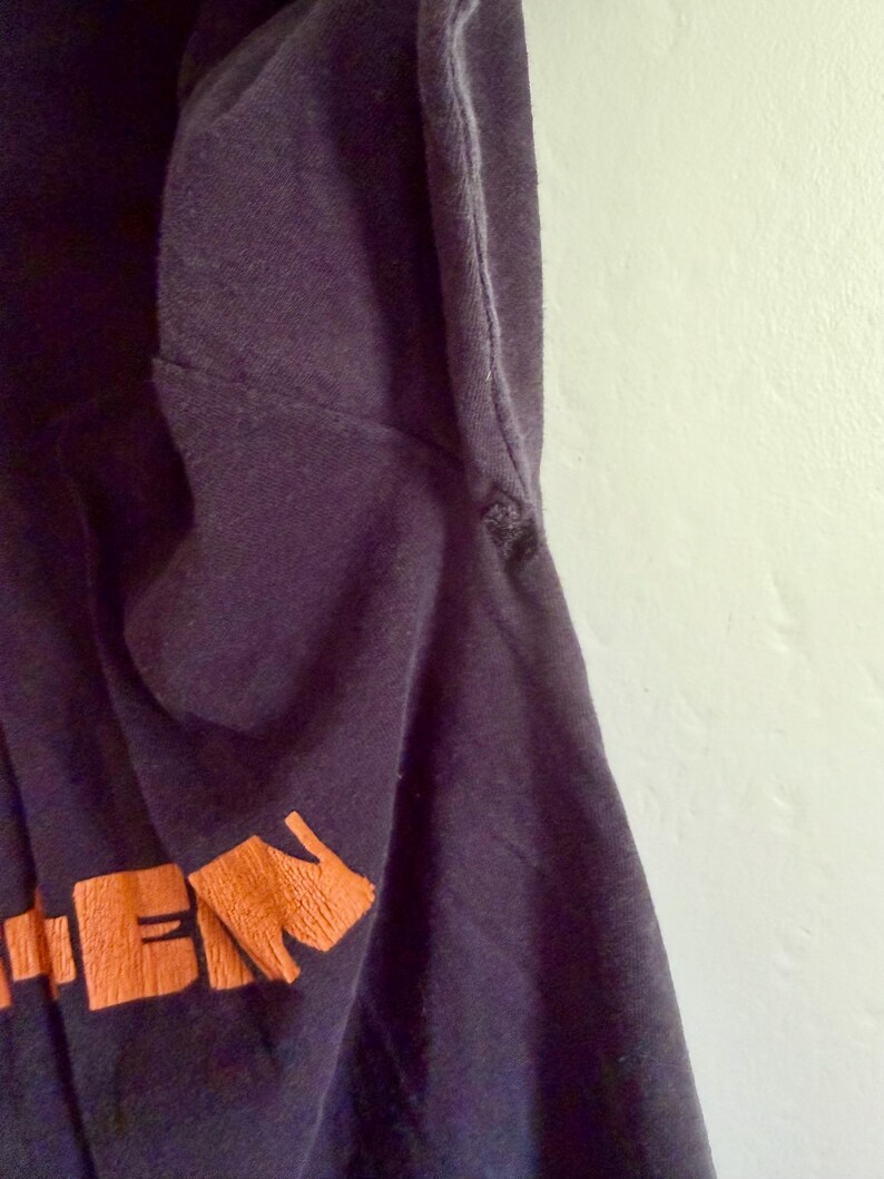 May include: A dark navy blue t-shirt featuring a portion of orange text printed on the lower section. The fabric shows a visible seam along the side and a small frayed area near the armhole, with a smooth texture throughout the garment.