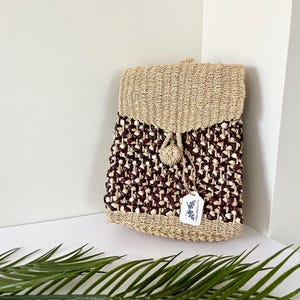 May include: A woven backpack with a flap closure. The top flap and bottom are a natural beige colour, while the body features a burgundy and beige pattern. A small beige pom-pom and a tag are attached. The bag is made of natural fibres.