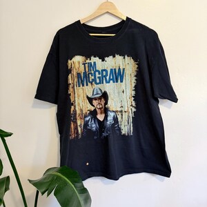 May include: Black t-shirt with a graphic of Tim McGraw wearing a cowboy hat and leather jacket. The text "TIM MCGRAW" is printed above the image in blue. The shirt is displayed on a wooden hanger.