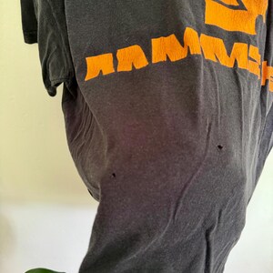 May include: A black short-sleeved t-shirt featuring the word RAMMSTEIN printed in bold orange letters across the chest. The fabric shows signs of wear, including two small holes visible on the front surface of the garment below the text.