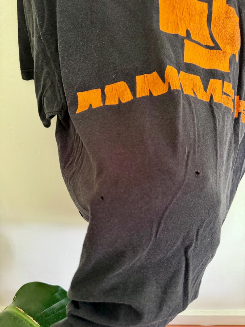 May include: A black short-sleeved t-shirt featuring the word RAMMSTEIN printed in bold orange letters across the chest. The fabric shows signs of wear, including two small holes visible on the front surface of the garment below the text.