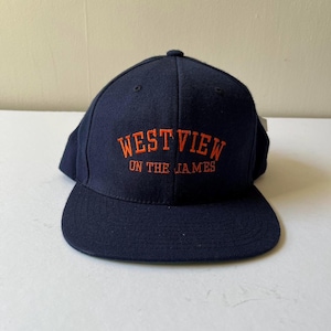 May include: A navy blue baseball cap with a flat brim. The cap features the embroidered text "WEST VIEW ON THE JAMES" in orange. The hat is made of a textured fabric and has a structured crown.