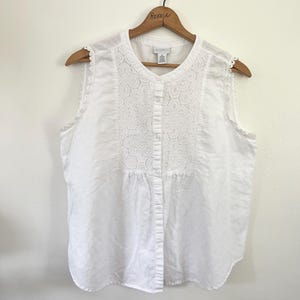 May include: White sleeveless blouse with a button-down front and a detailed lace panel on the bodice. The blouse has a rounded neckline and scalloped lace trim along the shoulders. It hangs on a wooden hanger.