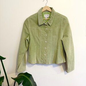 Peut inclure: Veste en daim vert clair avec un col et une fermeture boutonnée. La veste est suspendue à un cintre en bois contre un mur blanc. Manches longues et coupe ajustée.