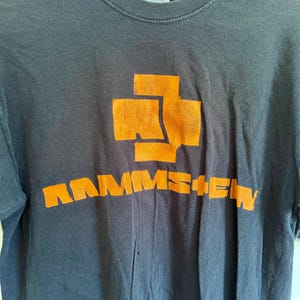 May include: A charcoal gray short-sleeved t-shirt featuring a large orange graphic on the chest. The graphic consists of a stylized cross symbol positioned above the text RAMMSTEIN in bold, blocky orange lettering. The fabric appears worn.
