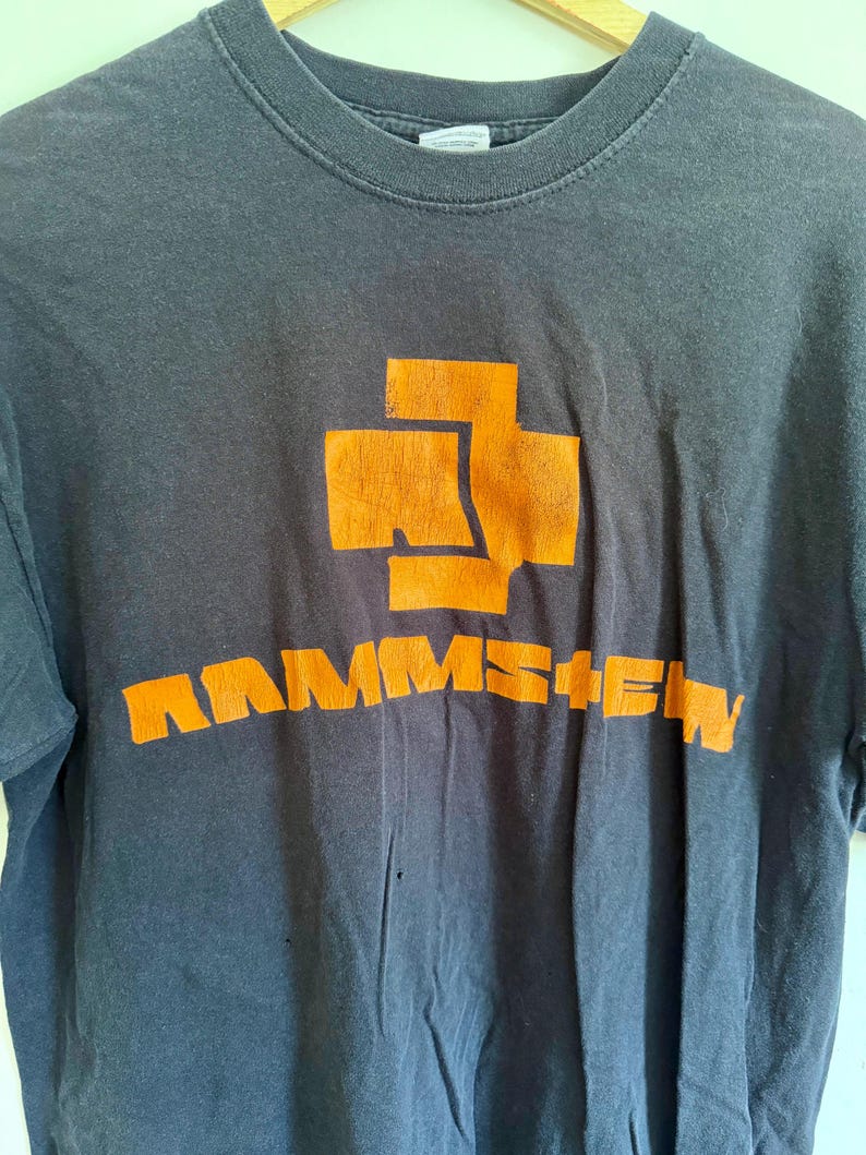 May include: A charcoal gray short-sleeved t-shirt featuring a large orange graphic on the chest. The graphic consists of a stylized cross symbol positioned above the text RAMMSTEIN in bold, blocky orange lettering. The fabric appears worn.