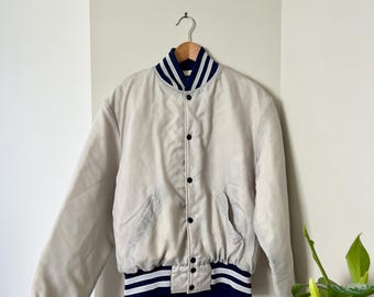 May include: A light beige varsity jacket with a navy blue and white striped collar, cuffs, and hem. The jacket has a button-up front and two front pockets. The jacket is hanging on a metal hook against a white wall.