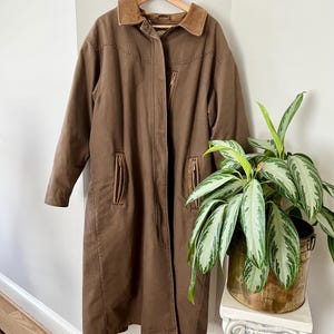 May include: A brown long coat with a corduroy collar and multiple pockets. The coat is hanging on a wooden hanger. The coat is a neutral color and appears to be made of a durable material.